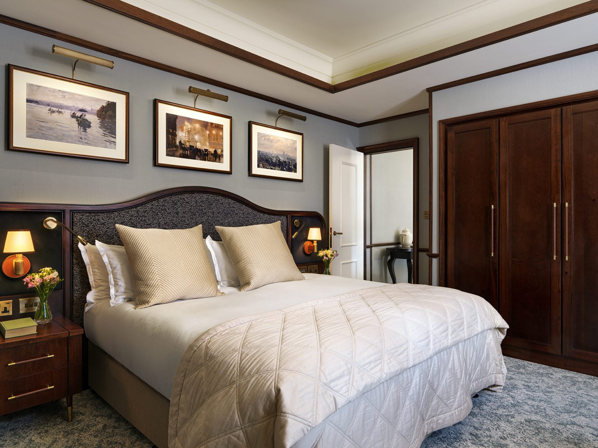 Experience the allure of a luxury five-star hotel suite in London. The elegantly appointed room features a large bed draped in a beige quilt, stylish wooden nightstands with lamps, and framed artwork above. A chic wooden wardrobe complements the décor next to a charming table by the open door.