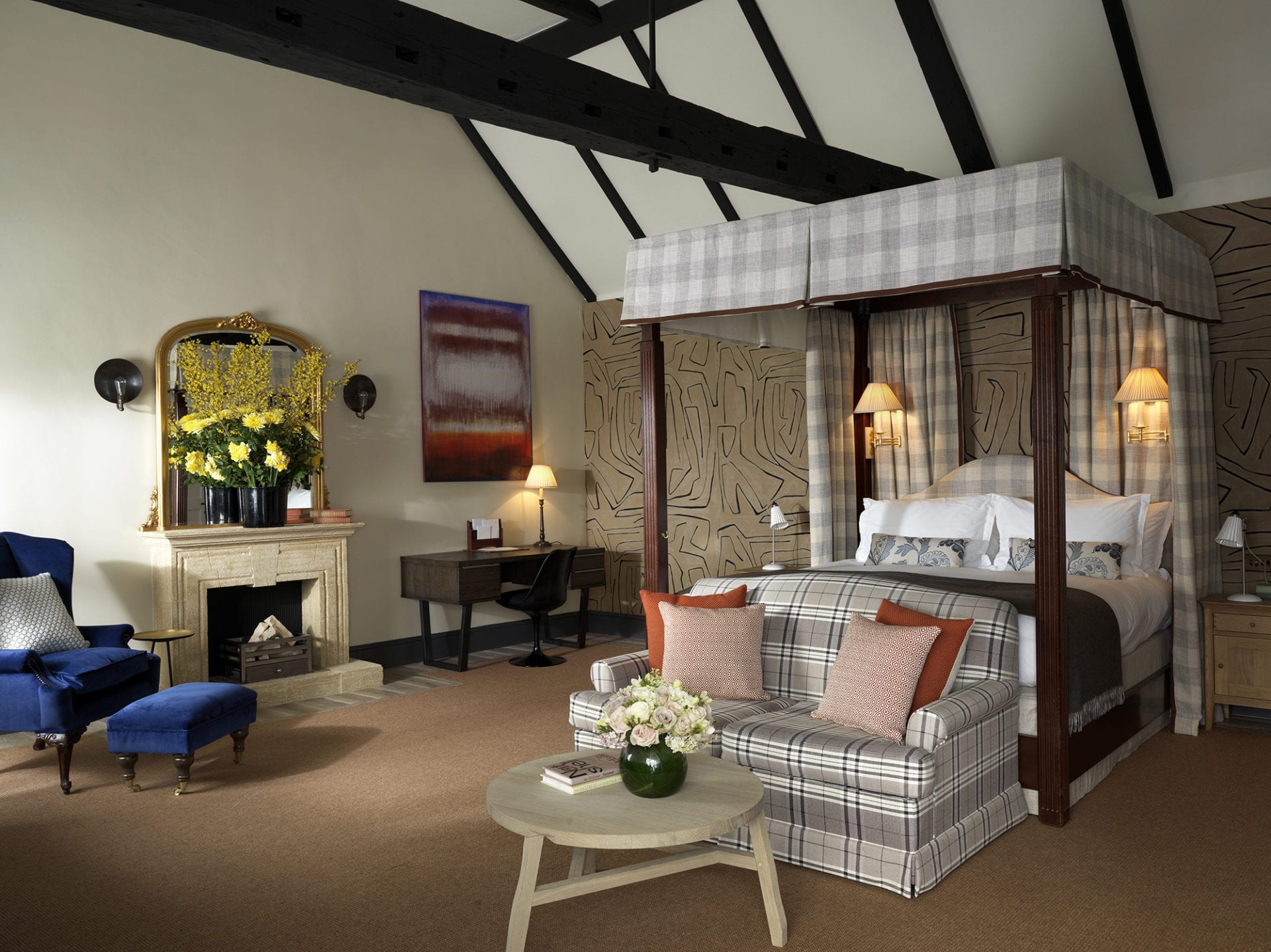 Luxurious bedroom with a four-poster bed, plaid sofa, and wooden table with flowers. A desk and chair are by the wall with abstract artwork. A fireplace with a mirror and yellow flowers is next to a blue armchair. Exposed beams adorn the ceiling.