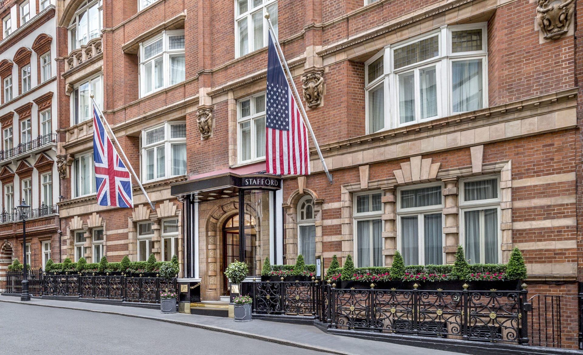 A red-brick hotel building with British and American flags at the entrance offers exceptional concierge & services, featuring black wrought-iron railings, potted plants, a large arched doorway, and several windows along a quiet city street.