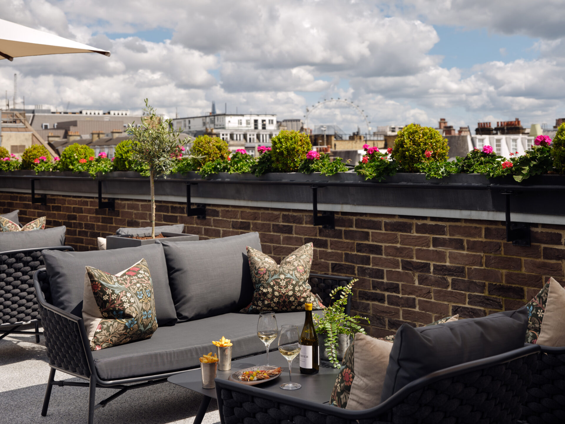 A rooftop patio at a luxury five-star hotel suite in London, featuring gray cushioned seating, patterned pillows, a table with wine and snacks, brick walls, potted flowers, and a city skyline under a partly cloudy sky.