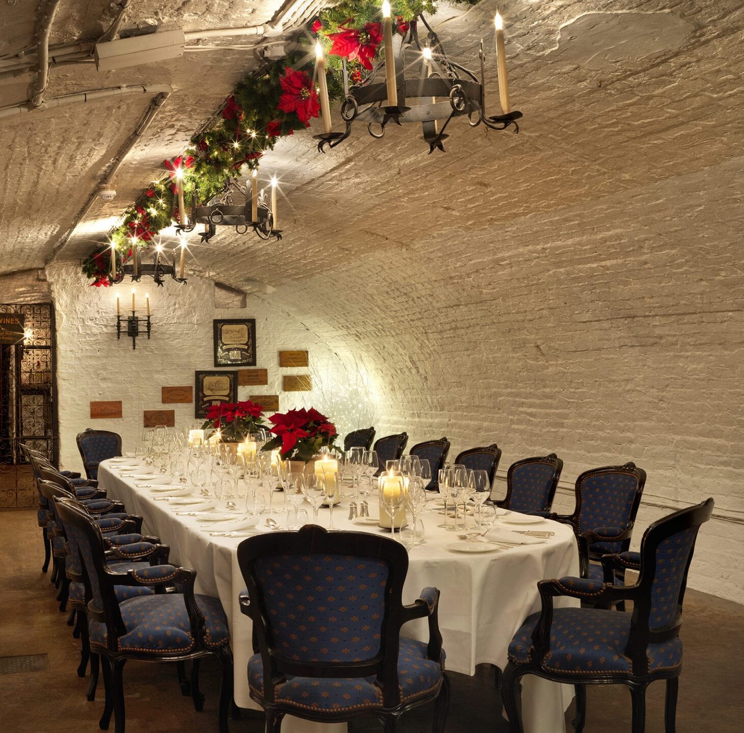 A long, elegant dining table with blue upholstered chairs is set for a formal meal in a brick-vaulted room. The table is decorated with red poinsettias, candles, and hanging garlands with lights and candles above.
