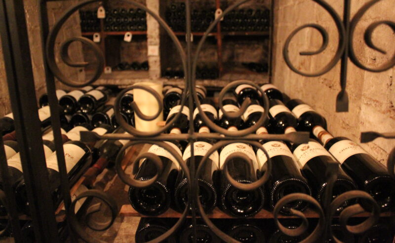 Bottles of wine are stacked horizontally on wooden racks in a dimly lit wine cellar, viewed through an ornate metal gate with swirling patterns. More wine bottles are visible on shelves in the background.