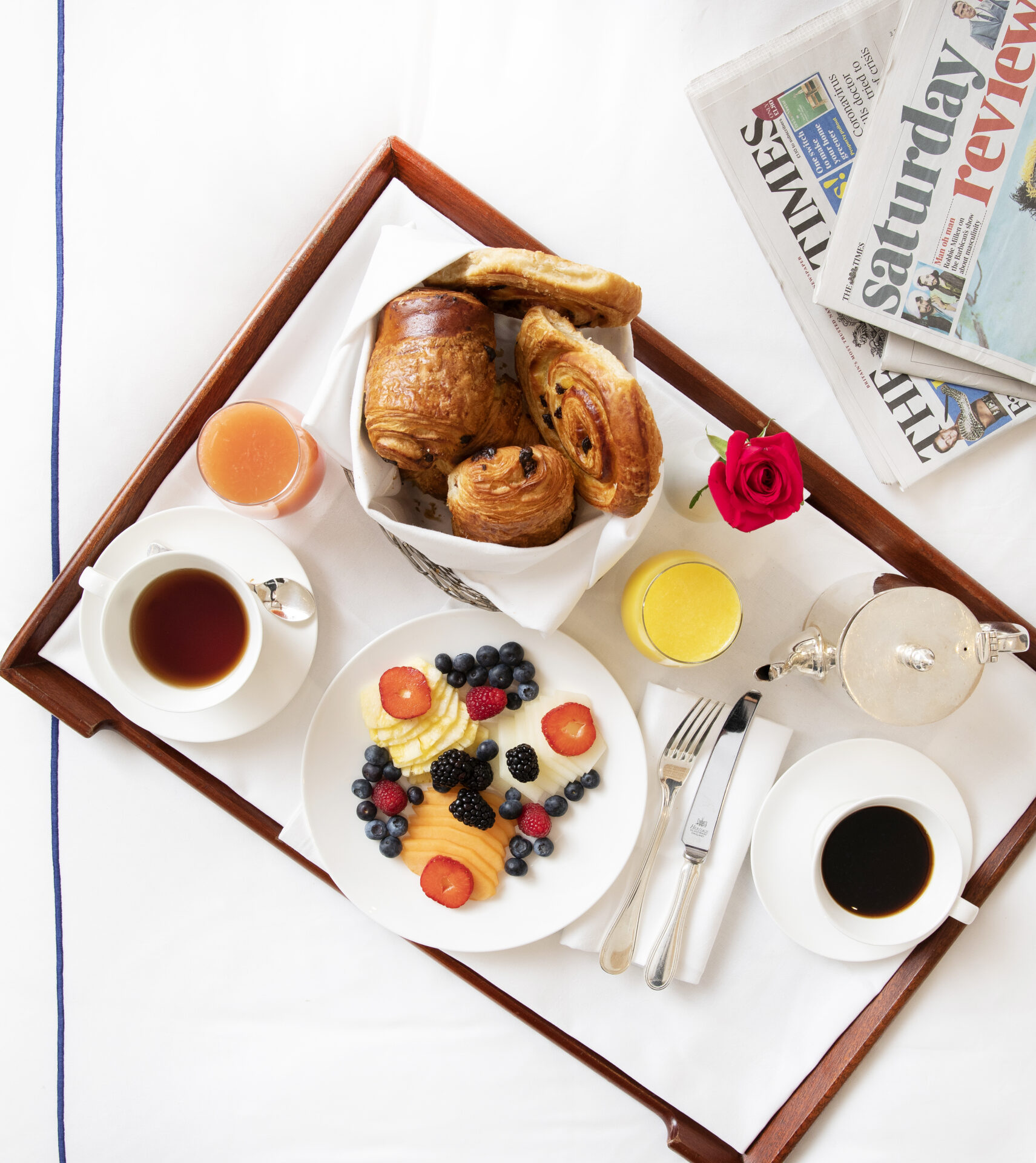 A luxurious breakfast tray on a bed at The Stafford London features a plate of berry-topped pancakes, two steaming cups of coffee, a glass each of orange and grapefruit juice, assorted pastries, a single red rose, and the morning newspapers for the perfect start to your day.