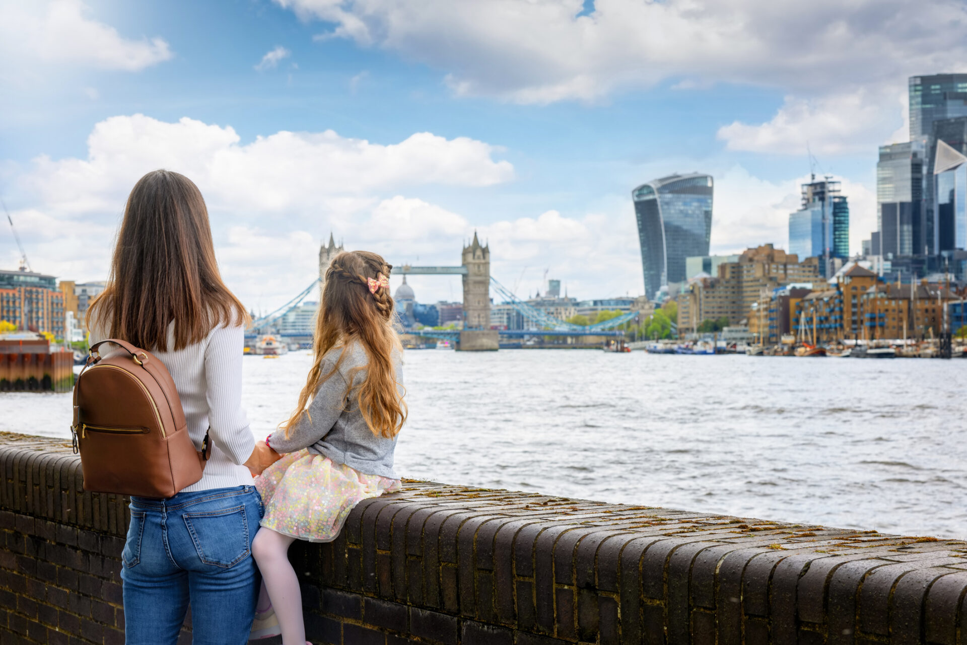 A woman with a brown backpack and a young girl sit on a brick wall overlooking the River Thames in London. Beyond, Tower Bridge and modern skyscrapers, including the Walkie Talkie building, rise against a cloudy sky. They discuss their London shopping adventure earlier around Mayfair.