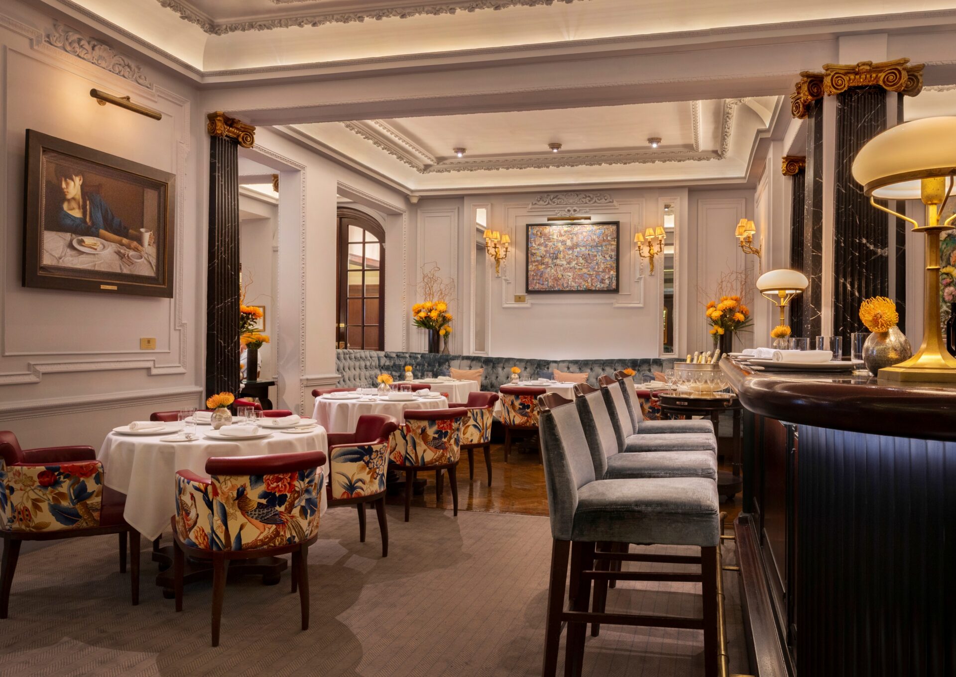 Elegant restaurant interior with gray tufted booth seating, floral-patterned chairs, and white marble tables. Walls are adorned with ornate moldings and a colorful abstract painting. Discover this refined ambiance alongside exclusive offers at The Stafford London. Warm lighting creates a cozy feel.