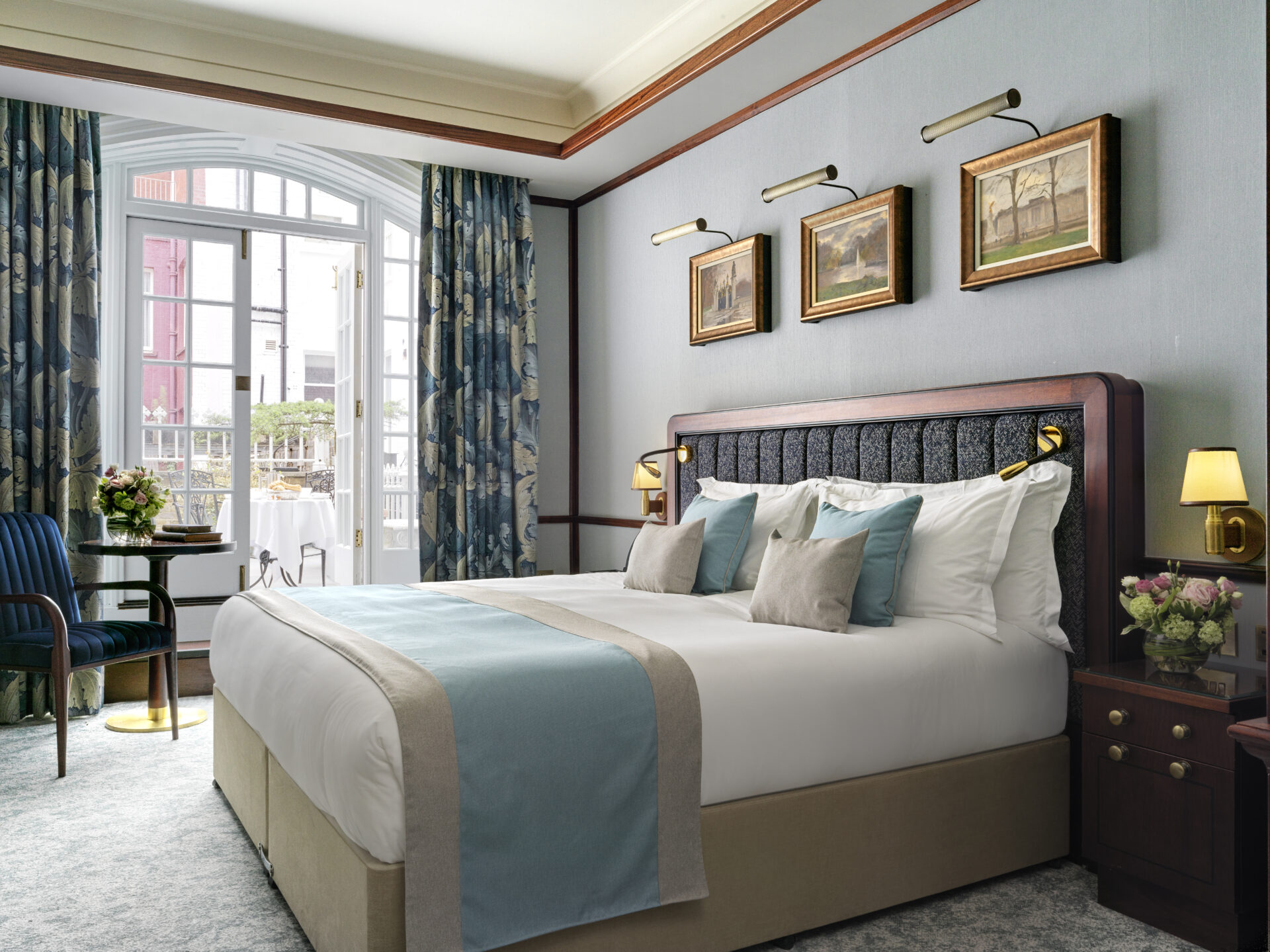Elegant bed and breakfast room with a large bed featuring blue and beige accents. Three paintings are above the headboard. The room has tall windows with patterned curtains, a small table with two chairs, and fresh flowers on bedside tables. Soft lighting adds warmth.