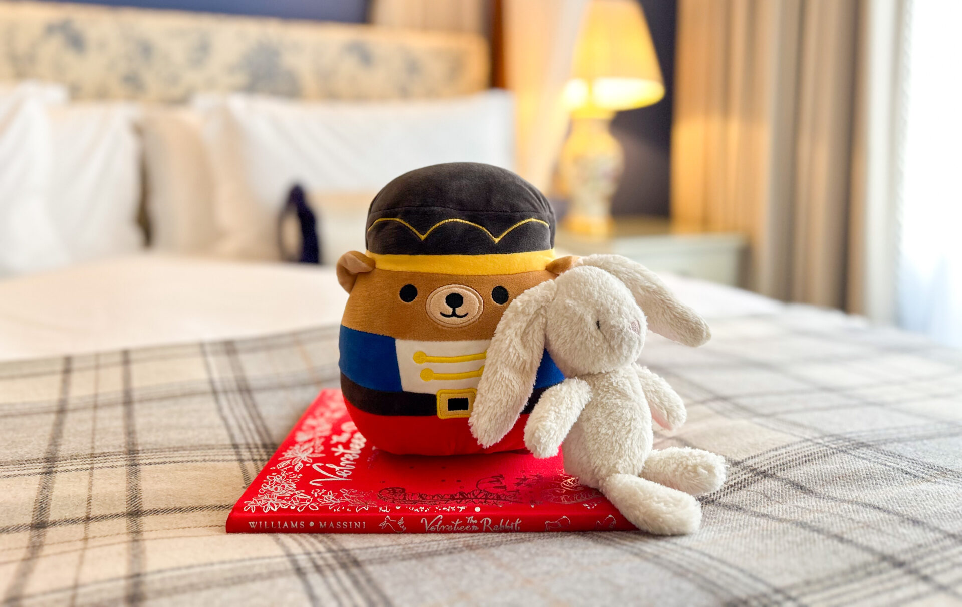 A plush bear dressed as a soldier and a white stuffed bunny sit on a red cloth atop a plaid blanket, forming an endearing family scene. In the background, a neatly made bed and softly lit lamp on the bedside table add to the cozy ambiance.