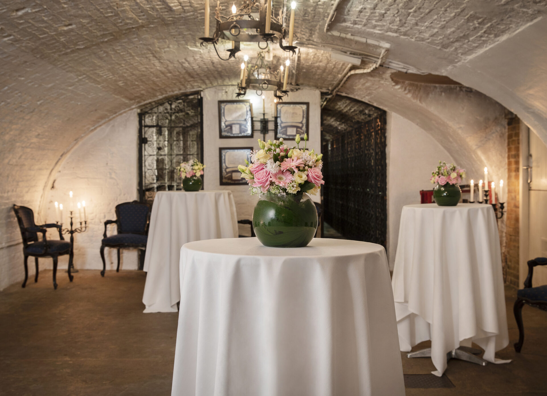 A dimly lit, elegant cellar with arched brick ceiling. Three tall tables covered in white cloth, each adorned with a vase of pink flowers. Chandeliers and candleholders provide soft lighting. Framed art and vintage chairs decorate the walls.