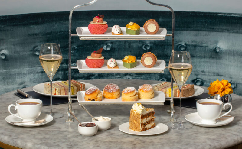 An elegant afternoon tea setup at The Stafford London features a three-tier stand with assorted pastries and desserts, two glasses of champagne, cups of tea, and a slice of cake on a marble table, set against a plush, dark green booth.