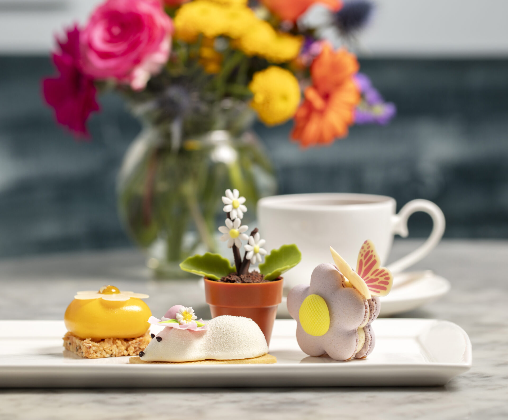 A plate of three intricate desserts, perfect for afternoon tea, including a yellow dome pastry, a white mousse dessert, and a purple flower-shaped treat with a butterfly decoration. A teacup and colorful flowers sit in the background.