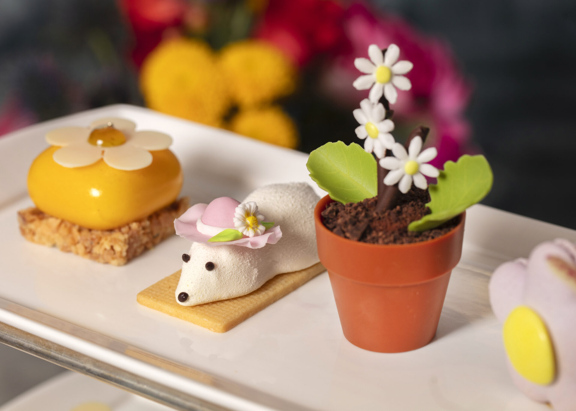 A plate with three desserts for afternoon tea: a flower-shaped cake, a white mouse-shaped treat with a pink hat, and a chocolate dessert in a small pot topped with edible flowers. Bright flowers blur in the background.