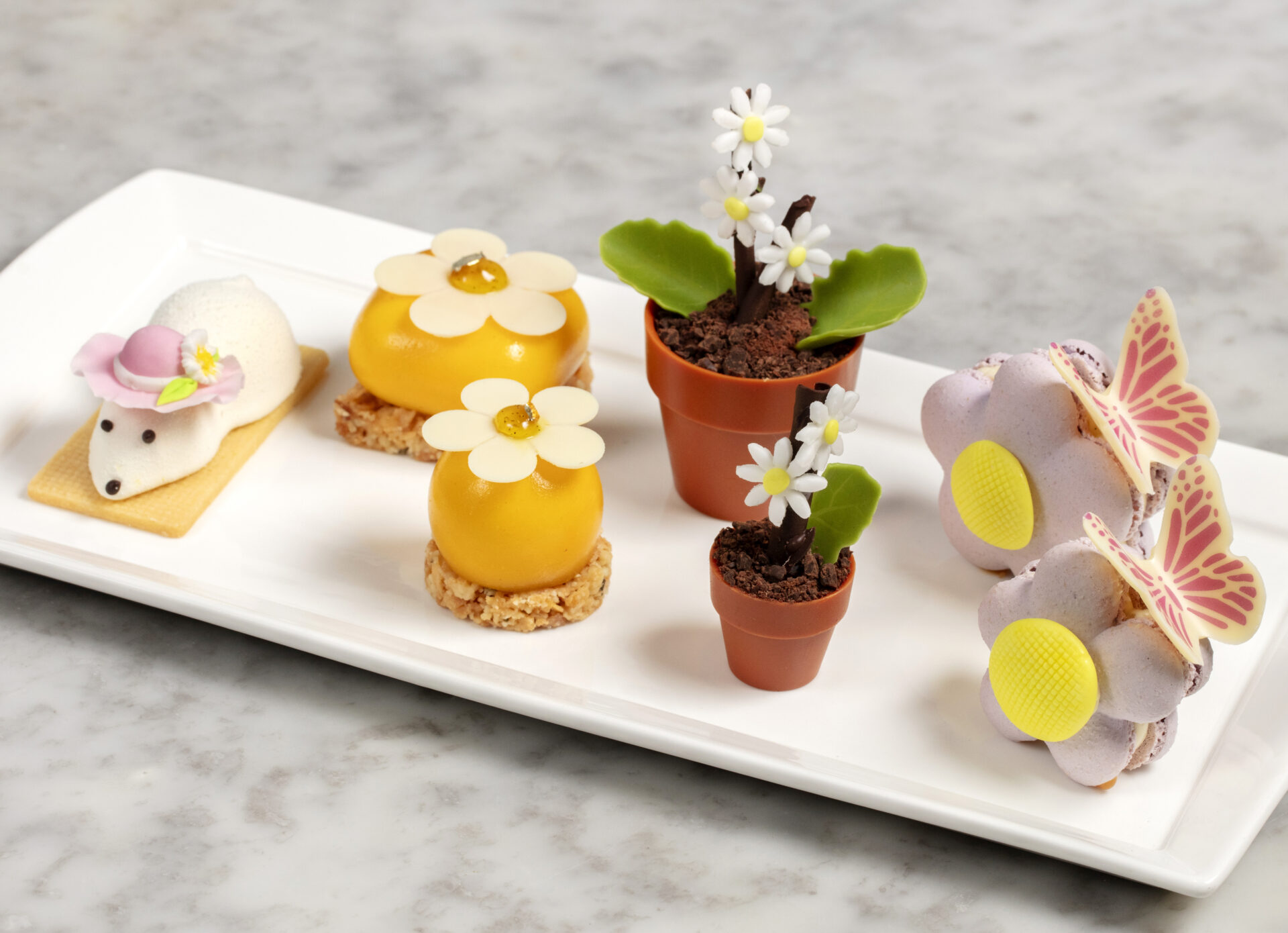 A white platter for afternoon tea displays desserts shaped like hedgehogs, yellow domes with white chocolate flowers, chocolate flower pots with daisies, and purple flower-shaped pastries crowned with butterflies.