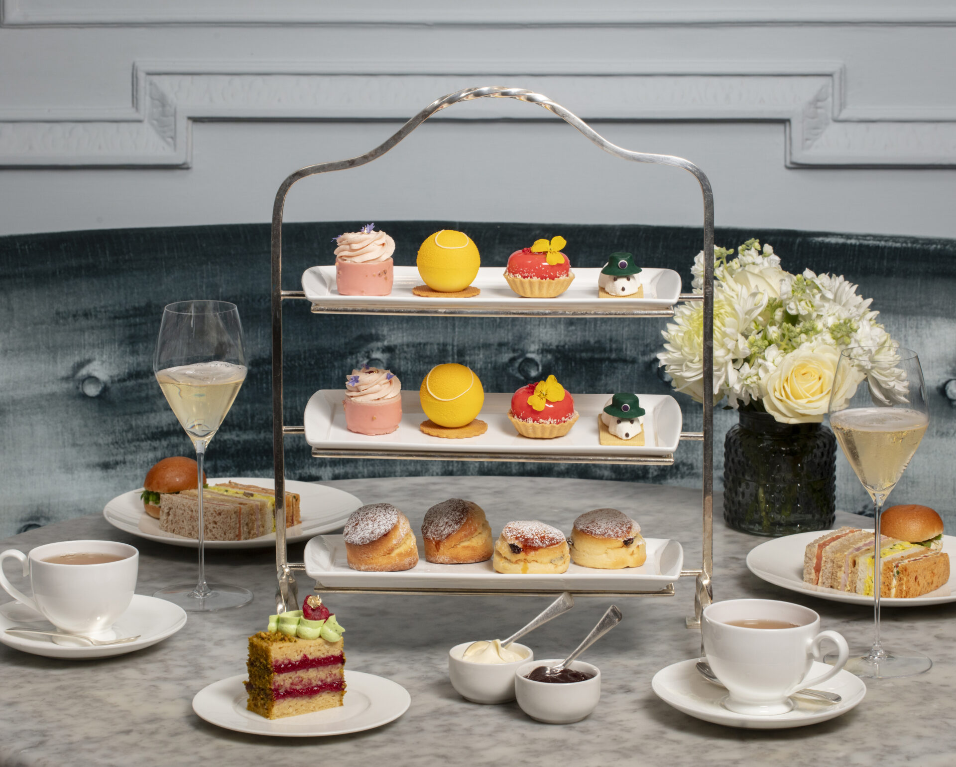 A three-tiered tray holds colorful pastries, scones, and cakes, surrounded by cups of tea, champagne glasses, finger sandwiches, and a vase of white flowers on a marble table—perfect for indulging in exclusive Dining Offers in an elegant setting.