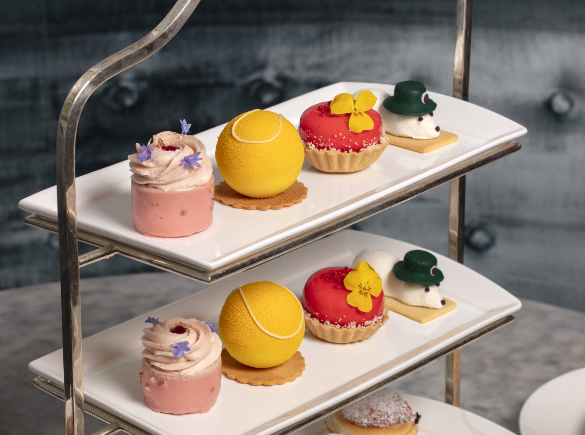 A two-tiered tray features elegant desserts as part of exclusive Dining Offers: yellow tennis ball-shaped pastries, round red tarts with edible flowers, pink mousse cakes with purple flowers, and white cookies topped with green hats.