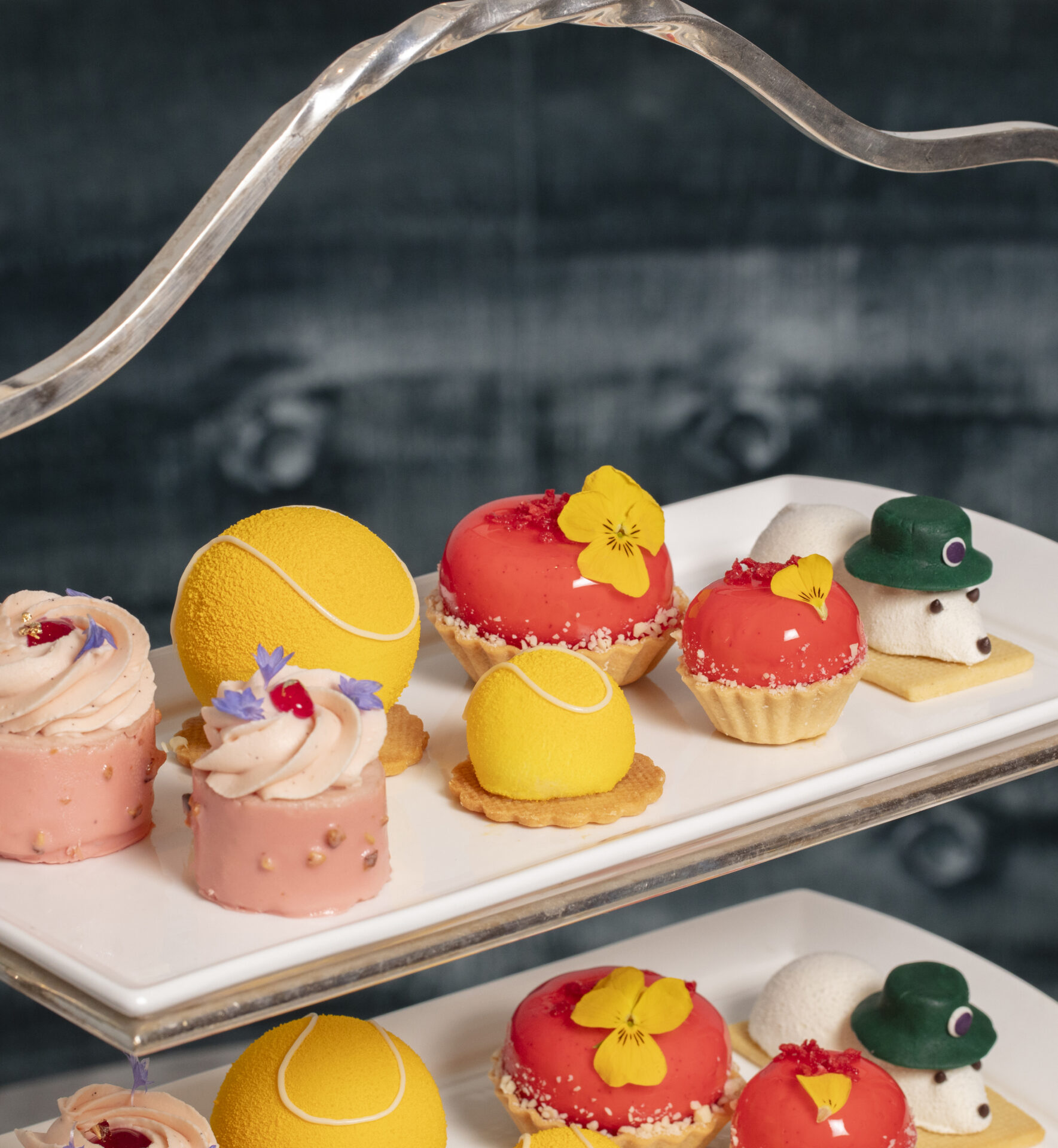 A tiered tray displays assorted colorful desserts, including yellow tennis ball-shaped cakes, red pastries with edible flowers, mini tarts, and white mousse shaped like mice with hats—perfect for indulging in delightful dining offers.