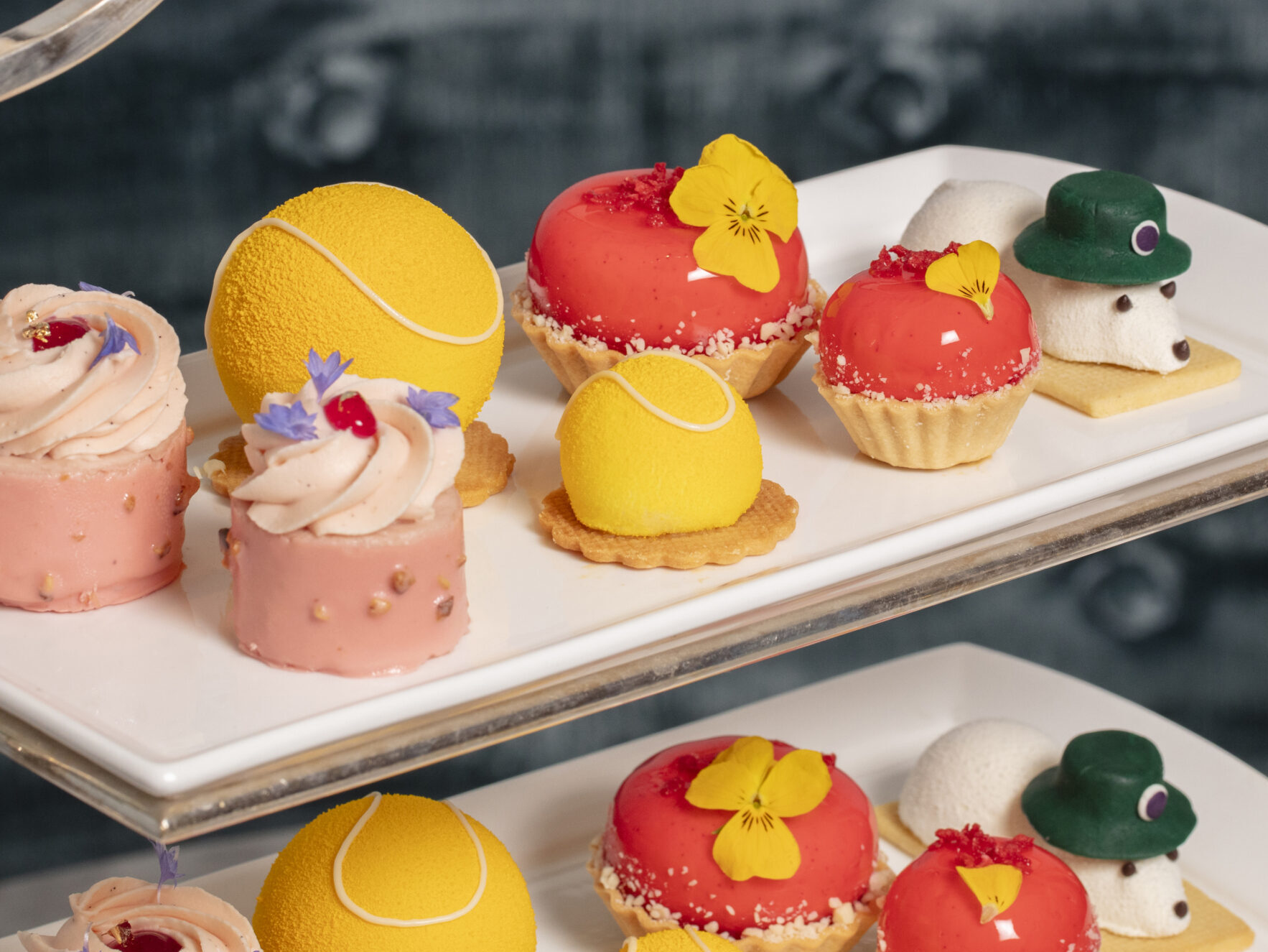 A tiered tray displays assorted colorful desserts, including yellow tennis ball-shaped cakes, red pastries with edible flowers, mini tarts, and white mousse shaped like mice with hats—perfect for indulging in delightful dining offers.