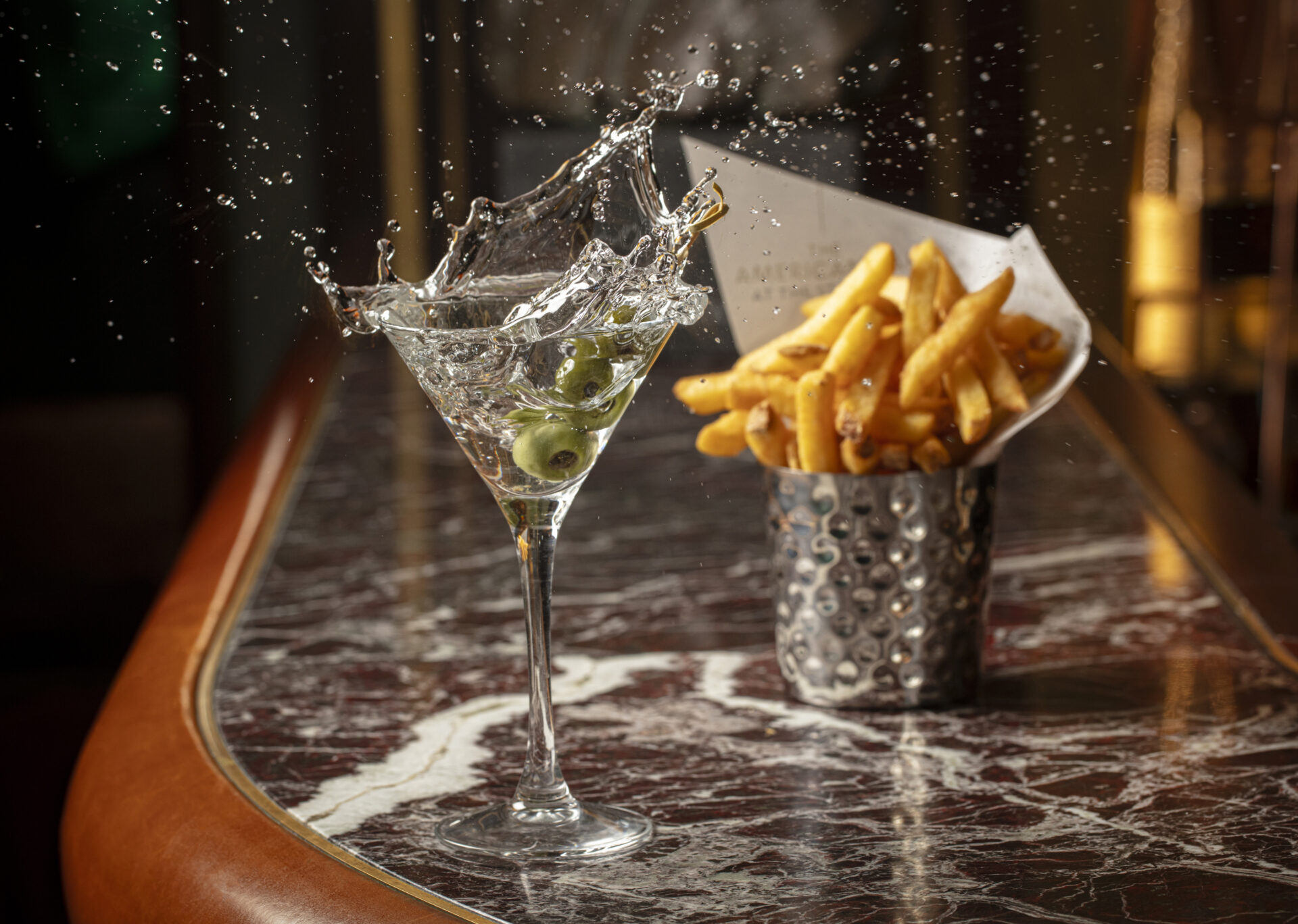 A martini glass with olives splashes as if just hit, sending droplets into the air. Beside it, a metal cup of French fries rests on a marble bar counter, hinting at tempting Dining Offers.