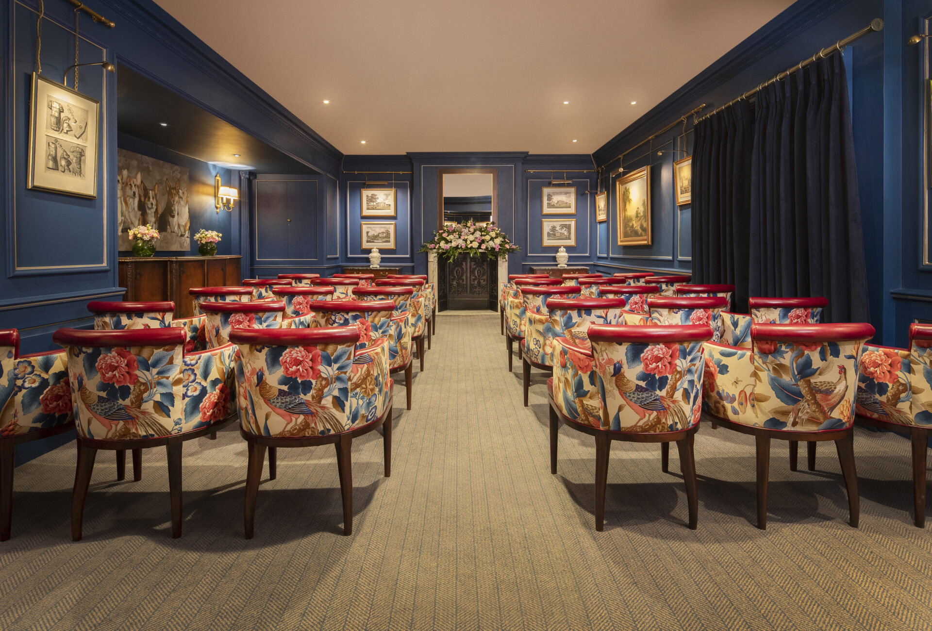 Rows of floral-patterned chairs face a flower-adorned podium in an elegant room with blue walls, framed artwork, and soft lighting, suggesting a setting for a ceremony or small event.