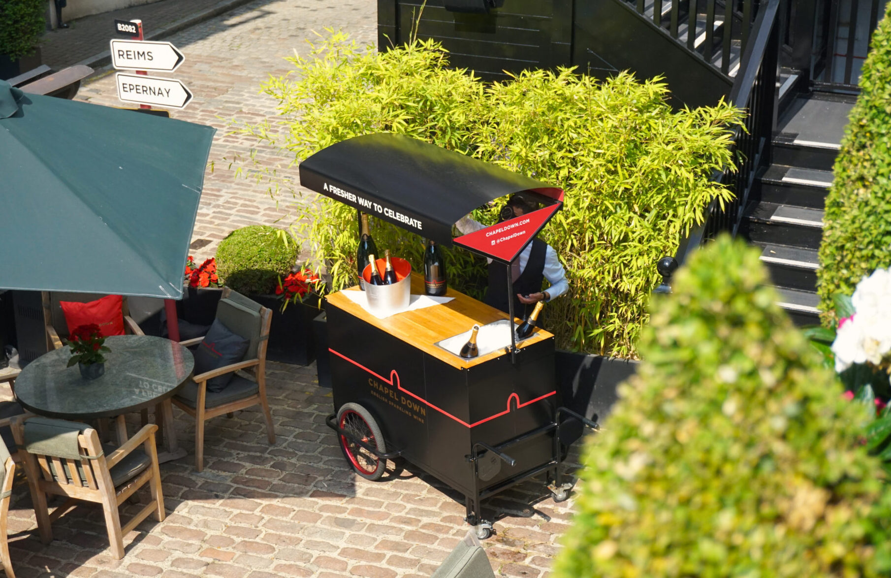 A black champagne cart with bottles in ice stands on a cobblestone patio near The American Bar, surrounded by green plants, outdoor seating with grey tables and chairs, and directional signs for Reims and Epernay.