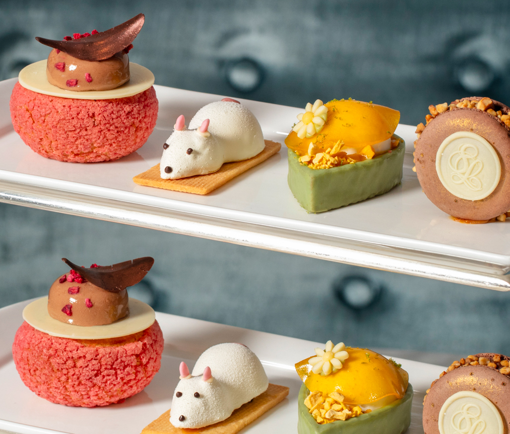A selection of elegant pastries for afternoon tea on white trays, including a pink cream puff, a white mouse-shaped dessert, a yellow fruit tart with green sides, and a chocolate mousse with a gold emblem.