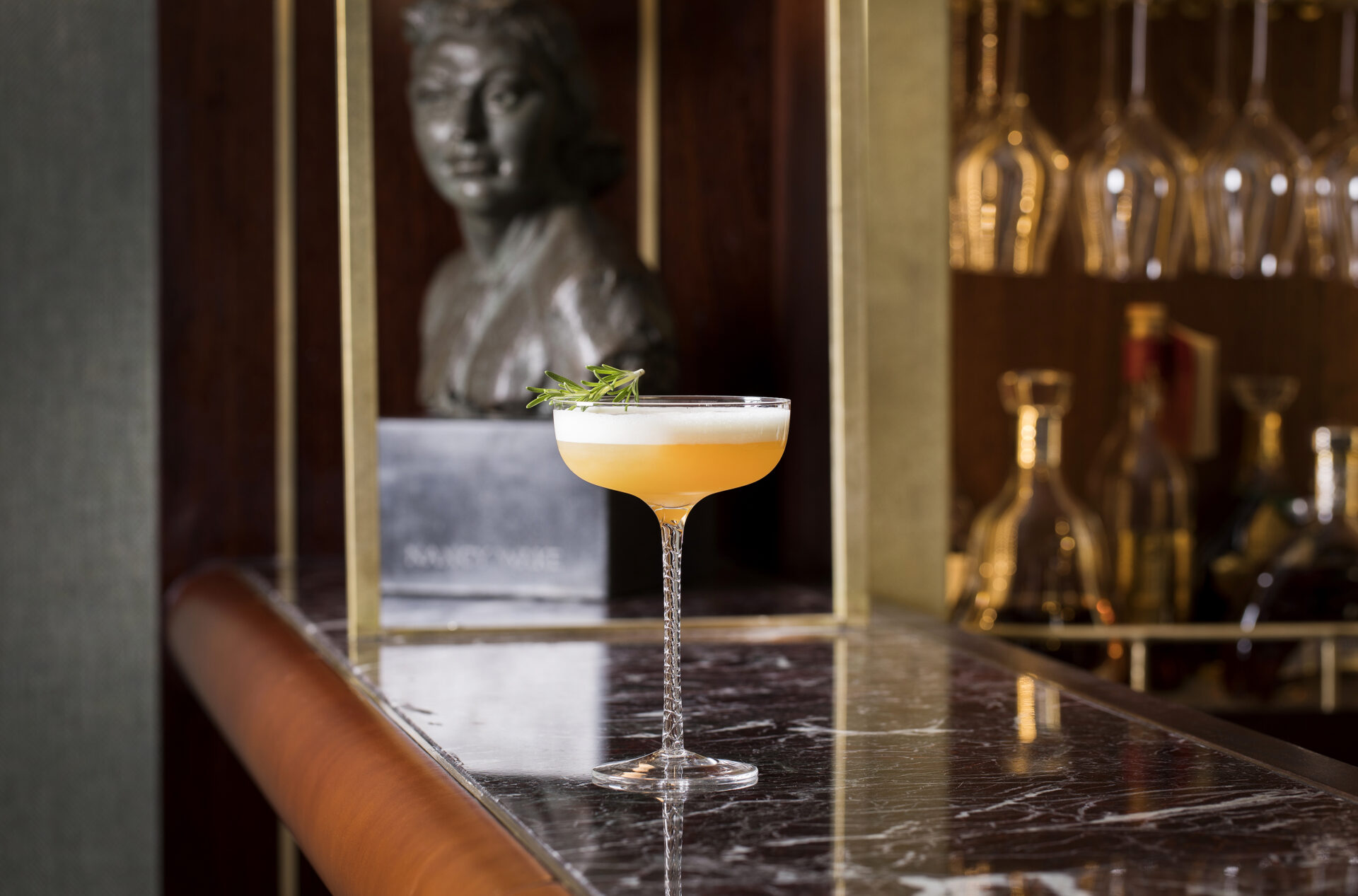 A yellow-orange cocktail with a foamy top and green garnish is served in an elegant coupe glass on a marble bar counter at Dining The American Bar. In the background, a bust sculpture and shelves with bottles and hanging glasses are visible.