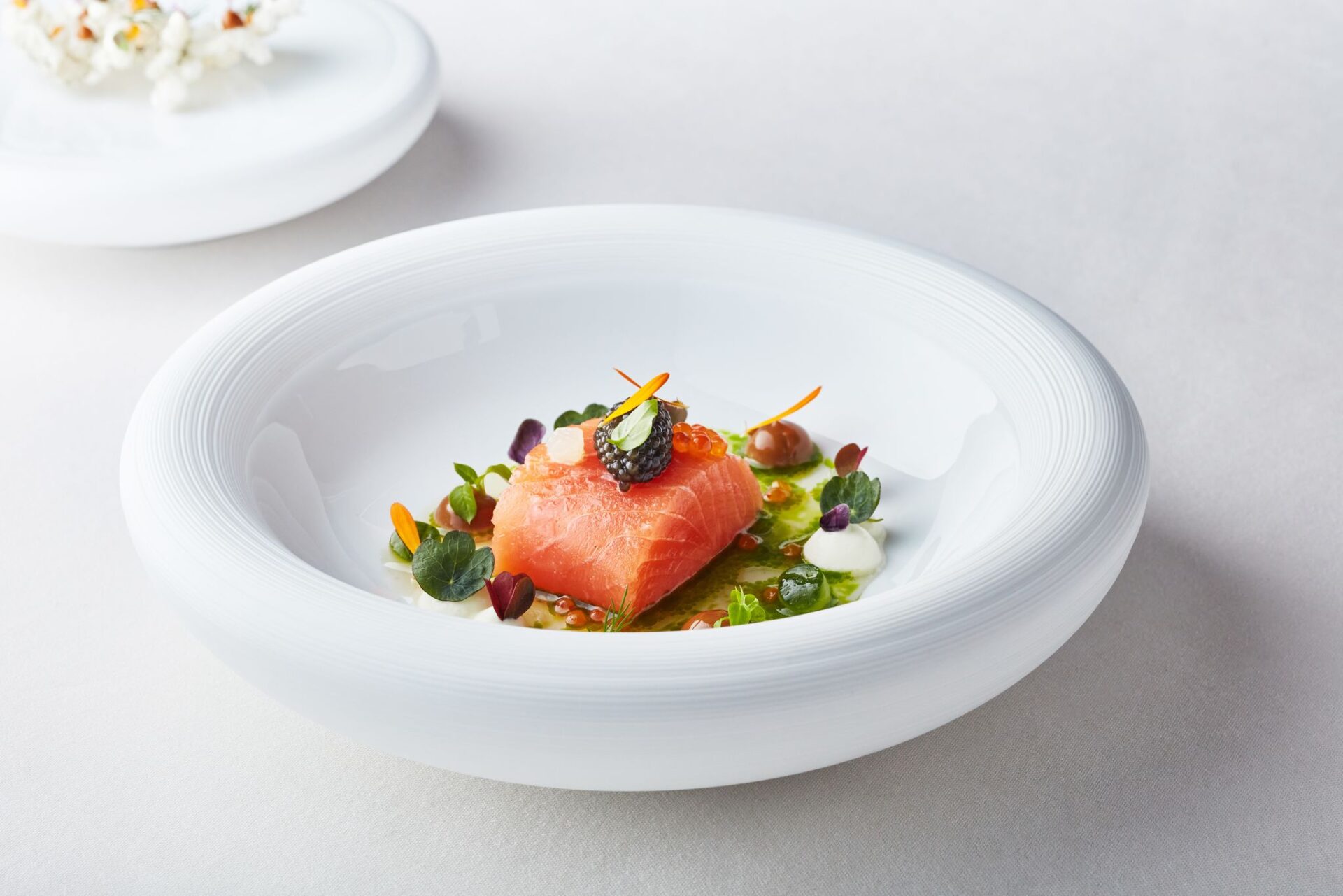 A gourmet dish featuring a slice of cured salmon topped with caviar, garnished with edible flowers, microgreens, and sauce, artistically presented on a white plate.