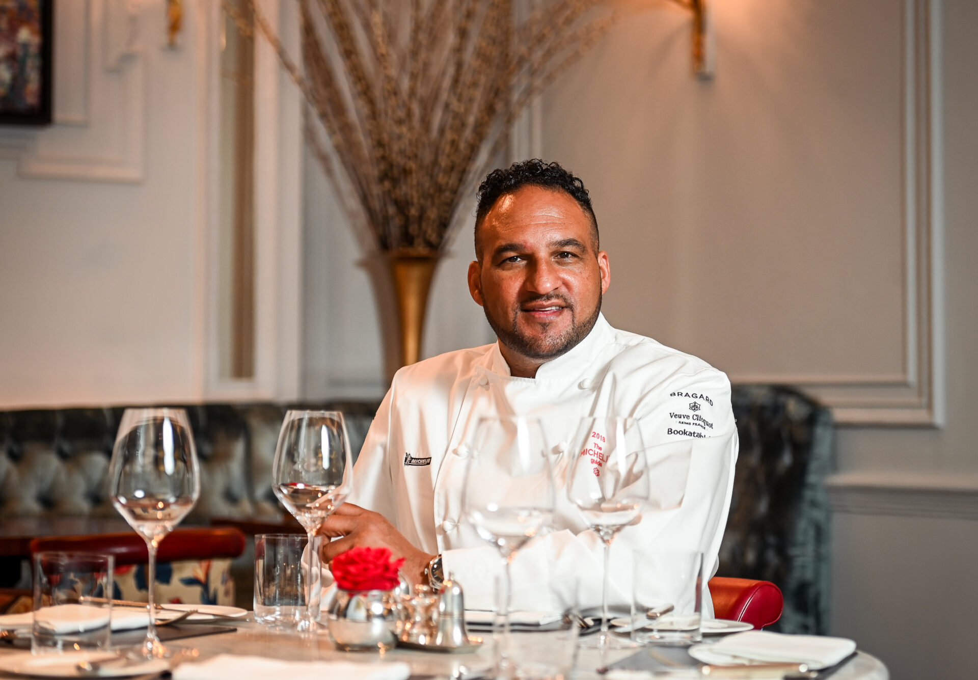 A chef in a white uniform sits at an elegant, set dining table with wine glasses and a red rose centerpiece, smiling in a warmly lit restaurant.