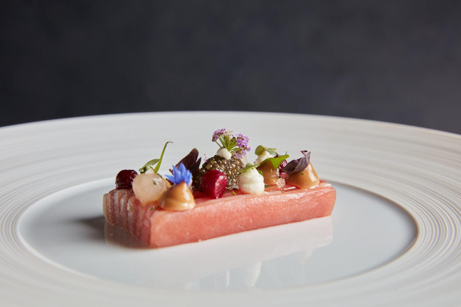 A rectangular slice of pink fish topped with small dollops of sauce, caviar, microgreens, and edible flowers, elegantly plated on a large white dish against a dark background.
