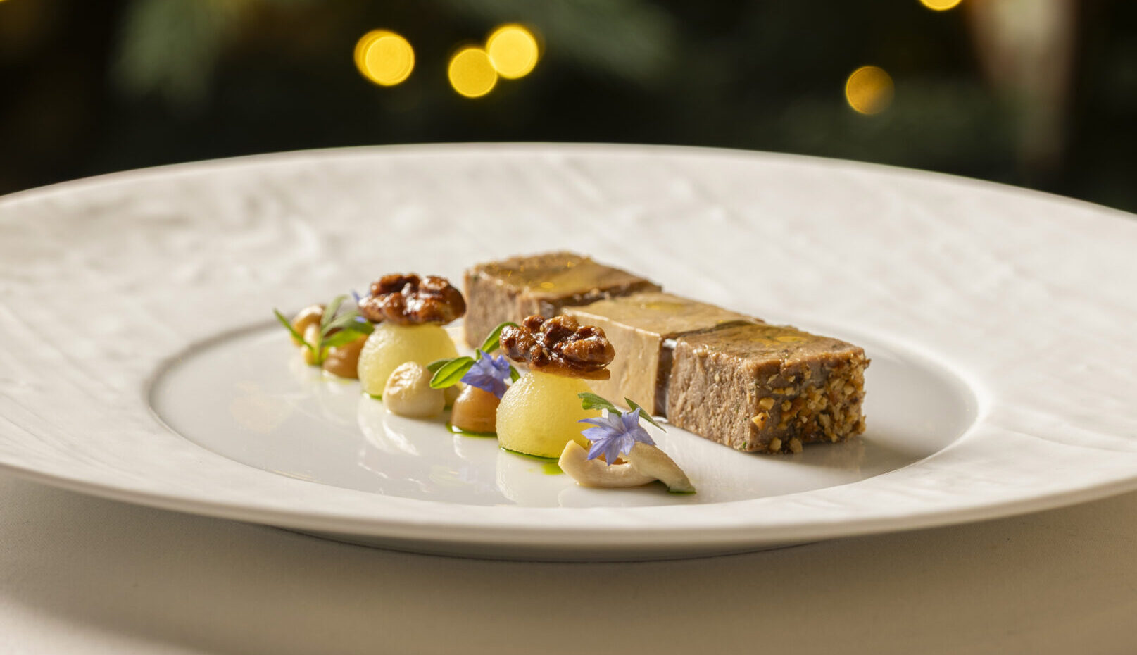 A gourmet dish from Michael Caines at The Stafford features two rectangular portions of pâté, garnished with nuts, herbs, and sauces on a white plate. In the background, blurred Christmas lights and ornaments decorate a tree.
