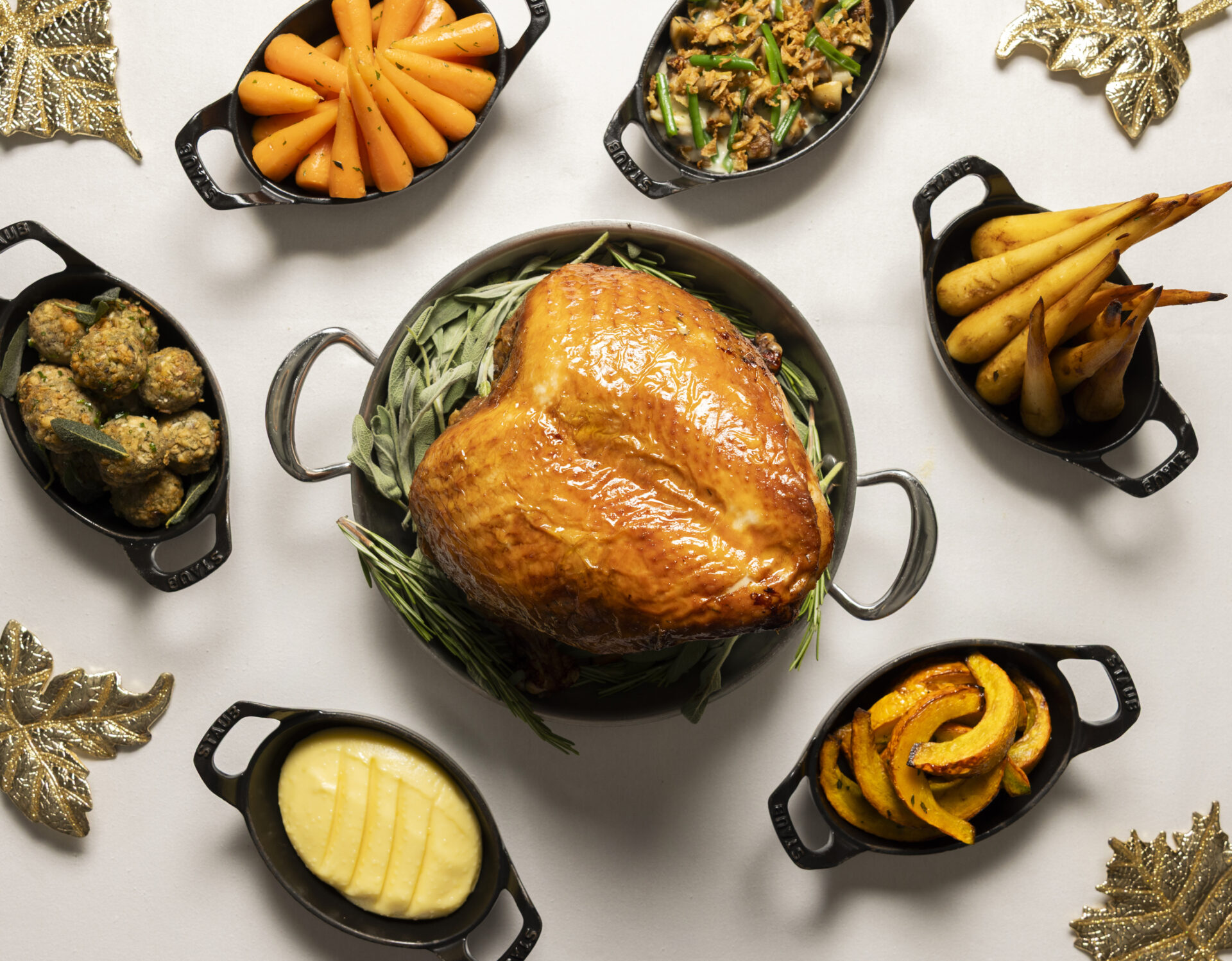 A roast turkey sits in the center surrounded by sides in small black dishes: carrots, green beans, roasted pears, squash, mashed potatoes, and stuffing balls, all arranged on a white table with gold leaf decorations.