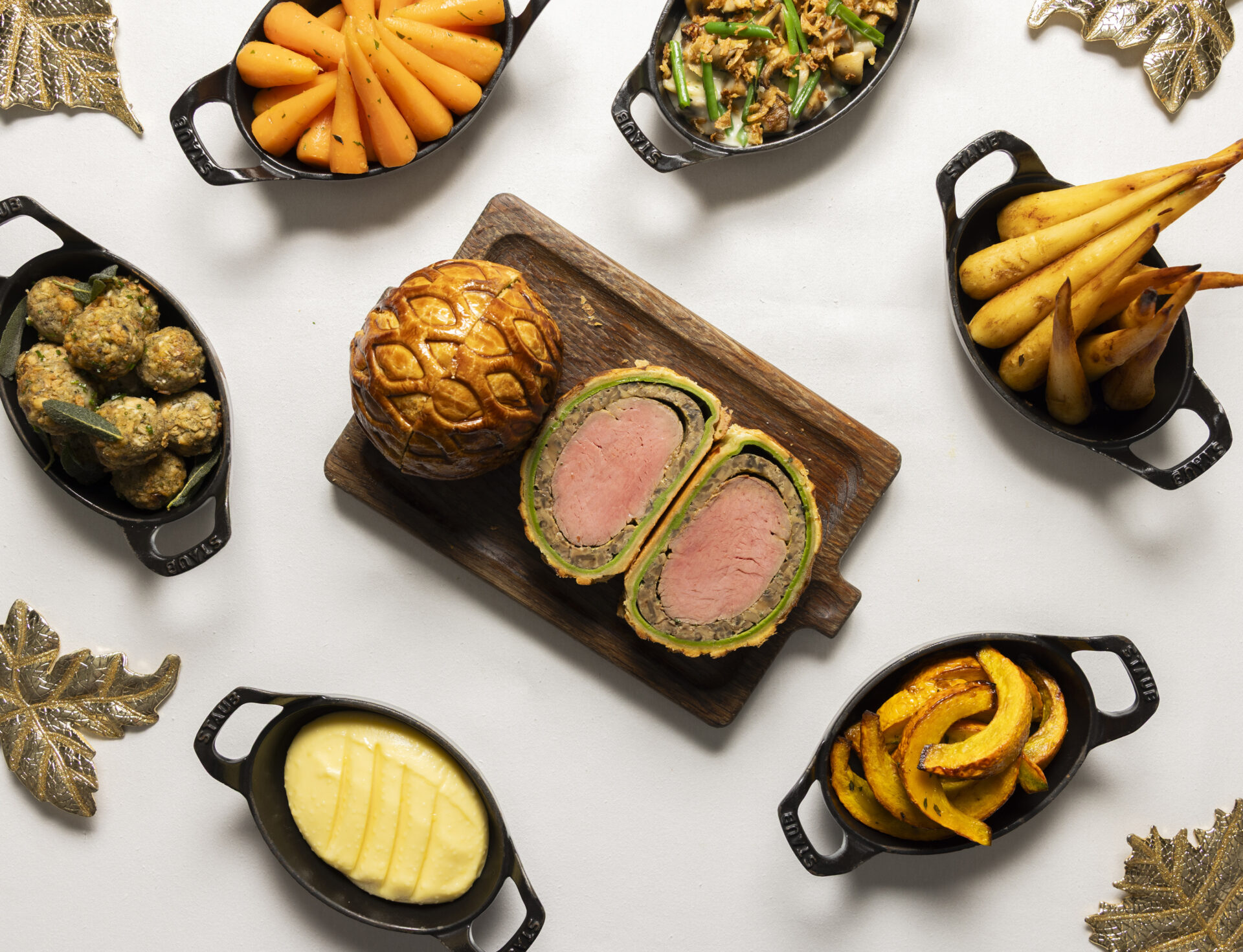 Overhead view of a wooden board with sliced beef Wellington and a pastry round, surrounded by trays of vegetables, potatoes, squash, carrots, and green beans on a white background with gold leaf decorations.