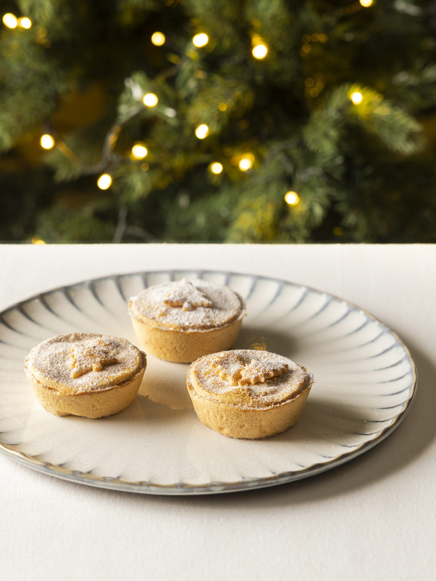 Three small mince pies dusted with powdered sugar sit on a decorative plate at Michael Caines at The Stafford, with a blurred background of a Christmas tree and glowing string lights.