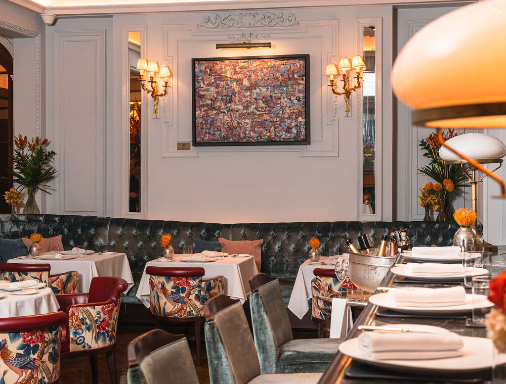 Elegant restaurant interior with plush velvet chairs, white tablecloths, and floral arrangements. Colorful wall art and soft sconce lighting create a warm, inviting atmosphere. Enjoy exclusive dining offers at the polished bar in the foreground.