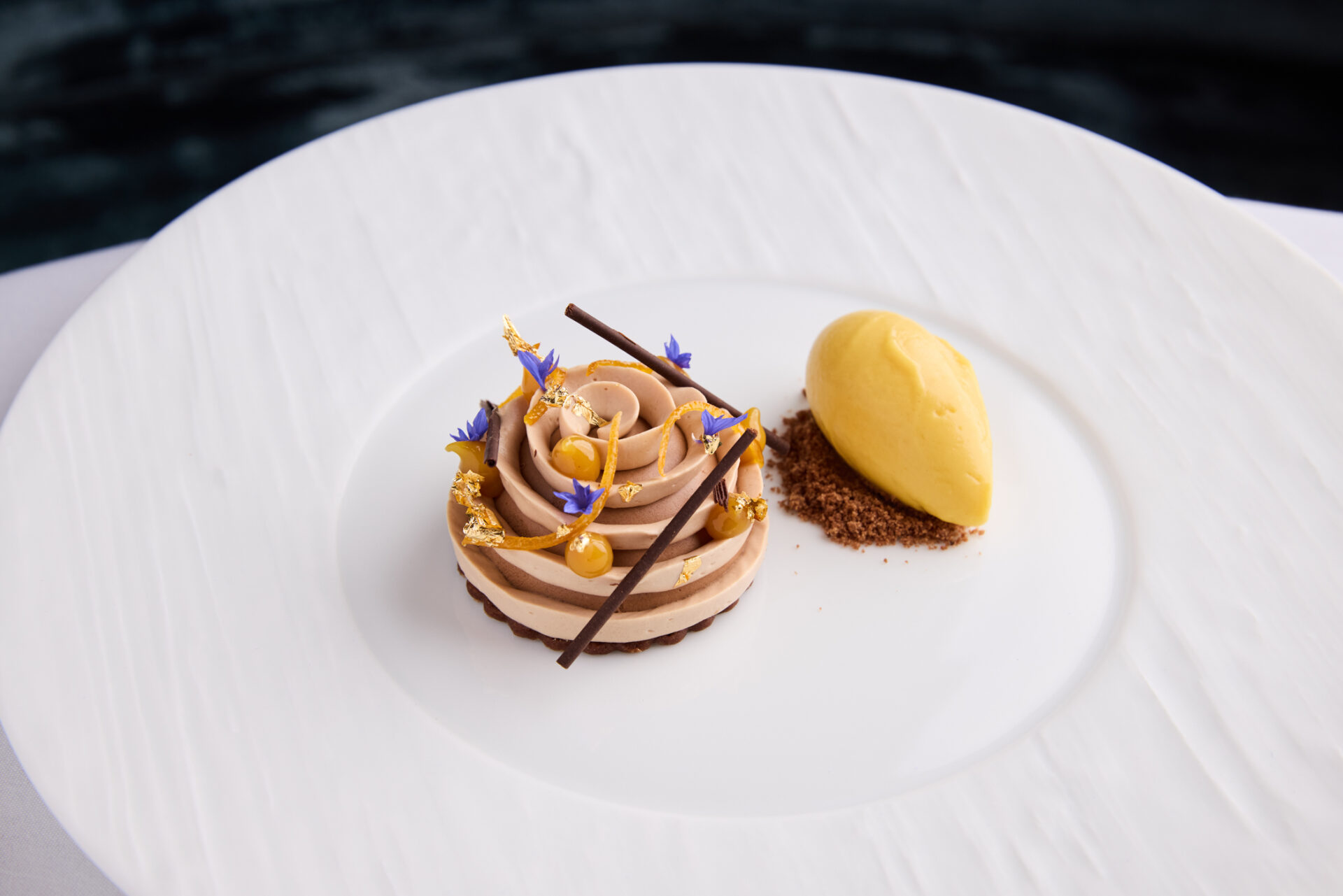 Elegant dessert at The Stafford London: chocolate mousse piped in a swirl, adorned with gold leaf, edible flowers, and chocolate sticks, served alongside a quenelle of yellow sorbet on a white textured plate.