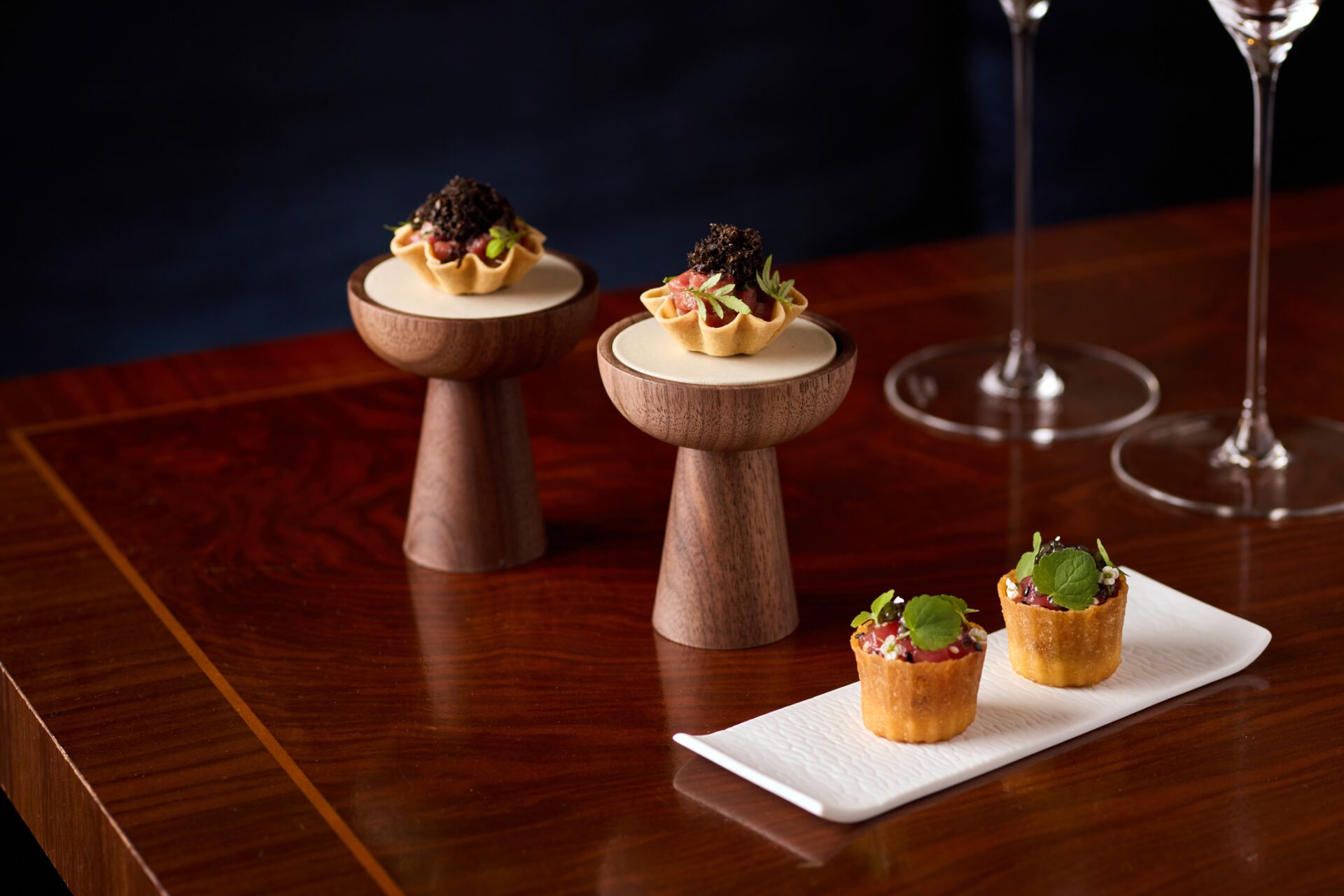 Two gourmet appetizers are displayed: two tartlets with dark toppings sit on wooden stands, and two small cup-shaped bites with green garnish rest on a white rectangular plate—perfect for sampling our exclusive Dining Offers.
