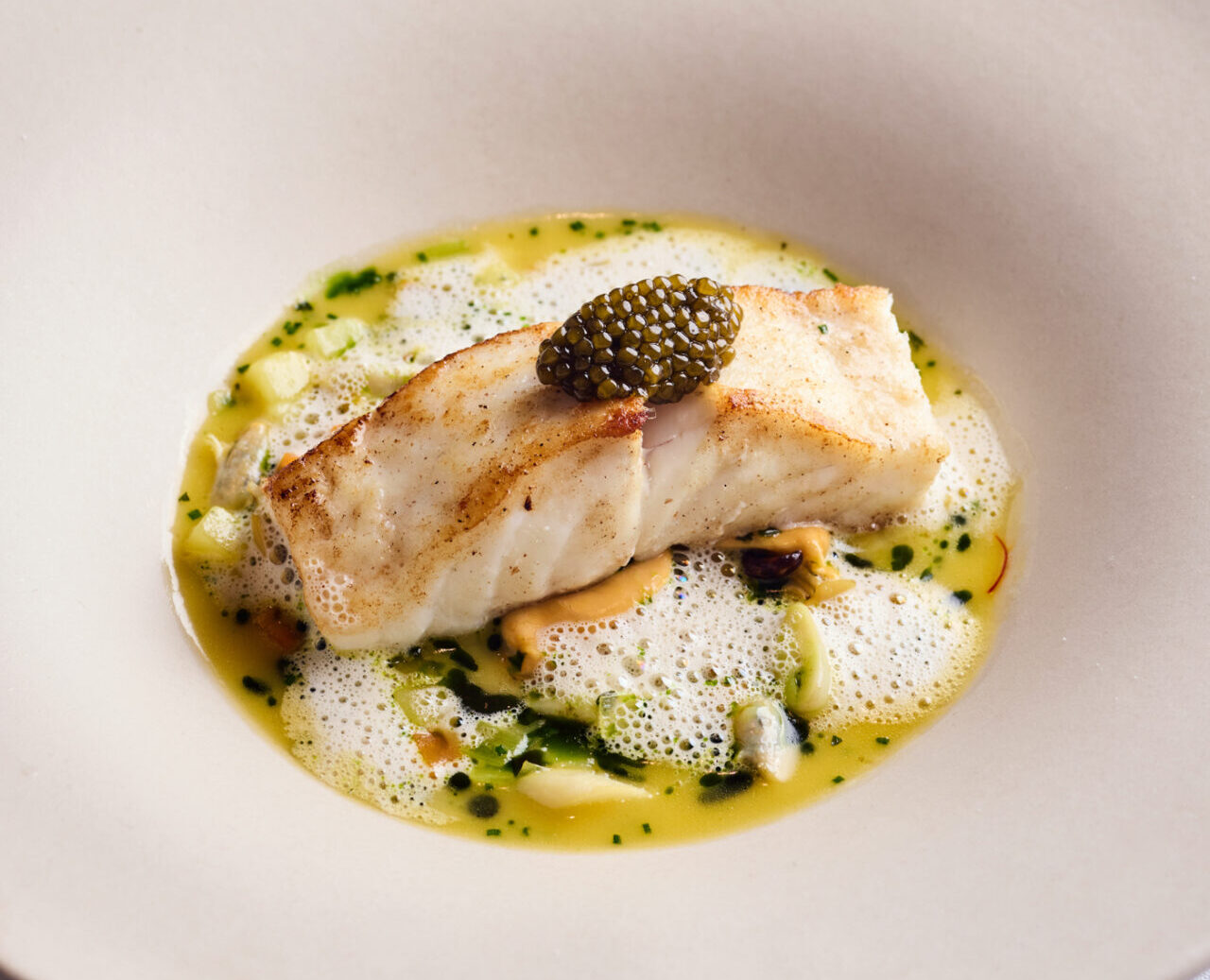 A fillet of cooked white fish topped with black caviar, served in a creamy yellow sauce with green herb oil, foam, and small vegetable pieces on a large white bowl-shaped plate—perfect for enjoying our exclusive dining offers.