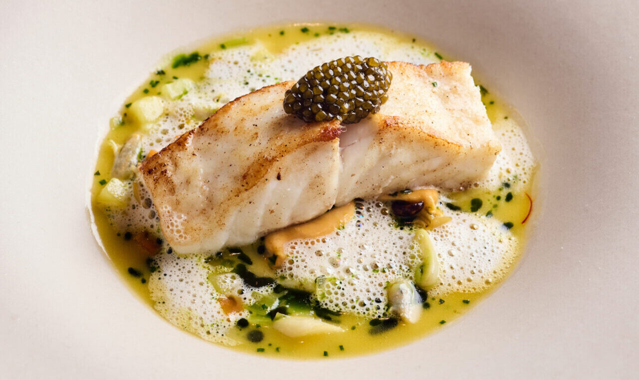 A fillet of cooked white fish topped with black caviar, served in a creamy yellow sauce with green herb oil, foam, and small vegetable pieces on a large white bowl-shaped plate—perfect for enjoying our exclusive dining offers.