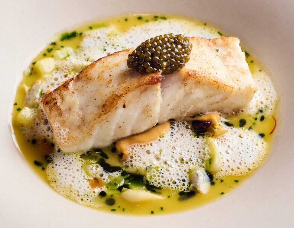 A fillet of cooked white fish topped with black caviar, served in a creamy yellow sauce with green herb oil, foam, and small vegetable pieces on a large white bowl-shaped plate—perfect for enjoying our exclusive dining offers.