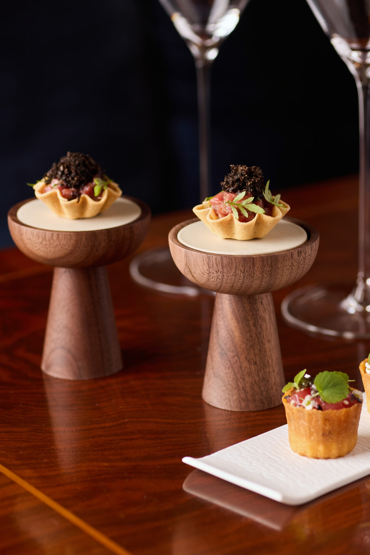 Two elegant bite-sized tartlets topped with shaved truffle and microgreens are displayed on wooden pedestal stands—perfect for Private Dining & Events, with two wine glasses and additional appetizers in the background.