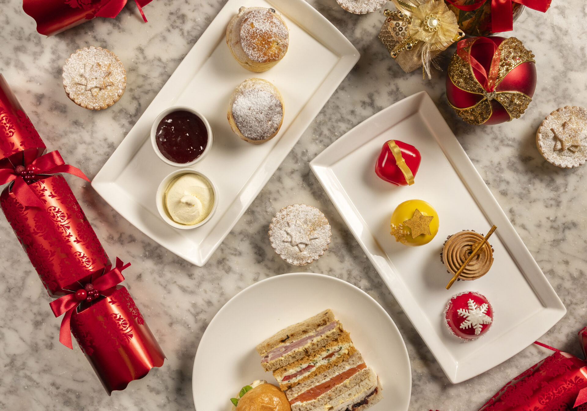 A festive table with Christmas crackers, decorated desserts, scones with jam and cream, mince pies, and a plate of assorted finger sandwiches, all arranged on a marble surface.