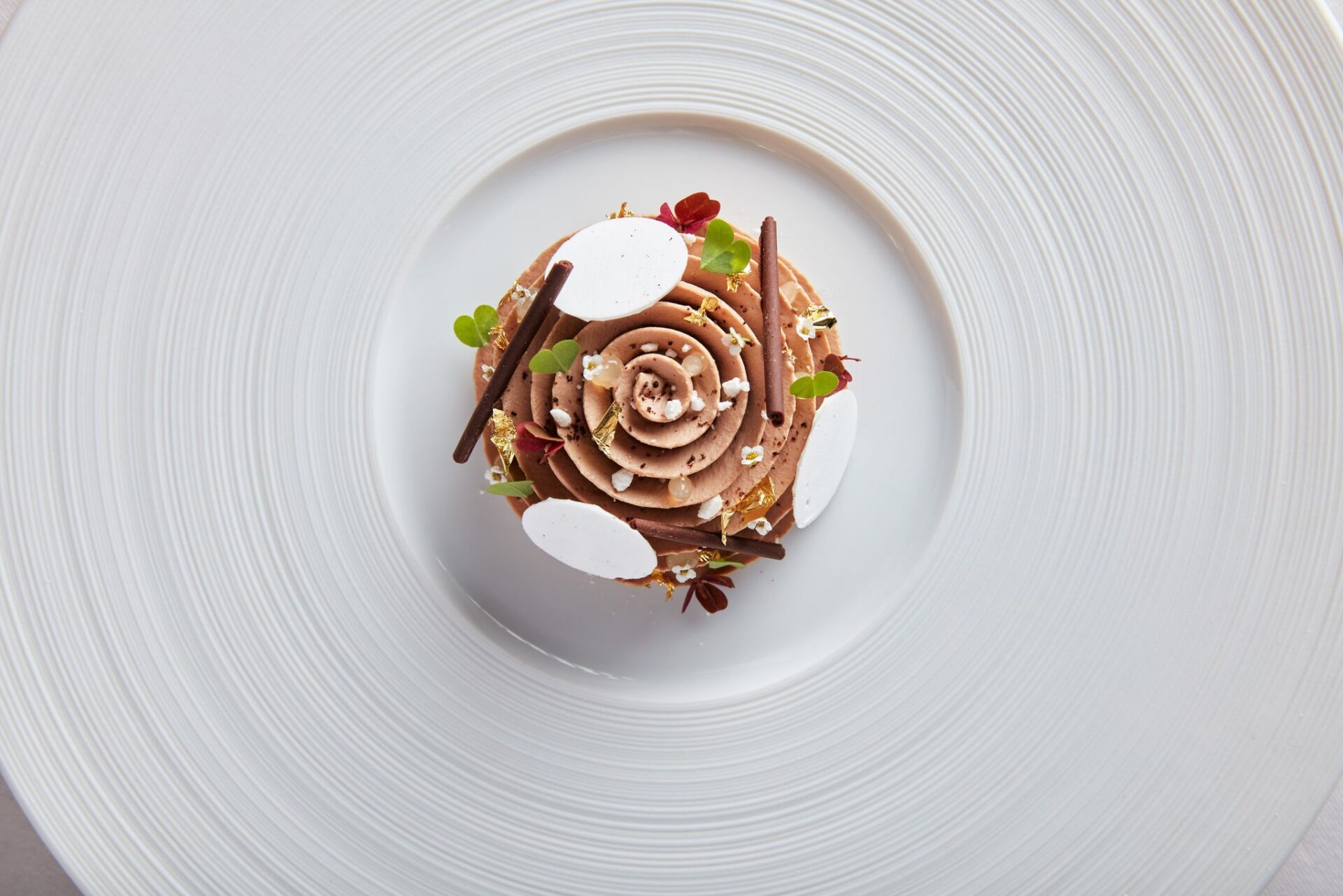 A beautifully plated dessert from The Game Bird features chocolate swirls, white meringue discs, chocolate sticks, and microgreens artfully arranged on a large white textured plate.
