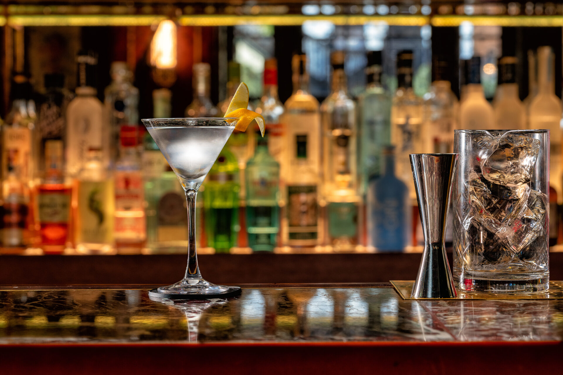 A martini with a lemon twist sits on a bar counter next to a glass filled with ice and a jigger, with blurred bottles of alcohol visible in the background.