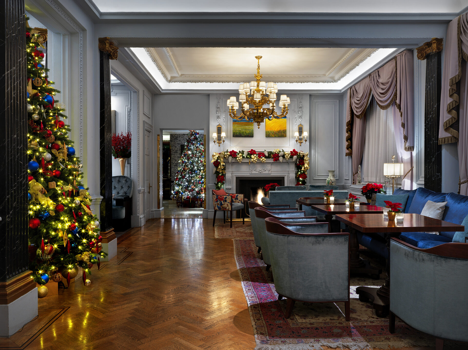 Elegant living room decorated for Christmas, featuring a large, lit Christmas tree with colorful ornaments, festive garlands on the fireplace mantel, blue velvet chairs, a wooden table, and soft, ambient lighting.