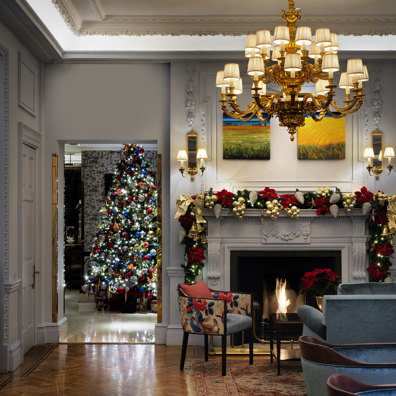 A festive living room decorated for Christmas, featuring a lit fireplace, elegant chandelier, garlands and red bows on the mantle, and a brightly lit Christmas tree in an adjoining room.