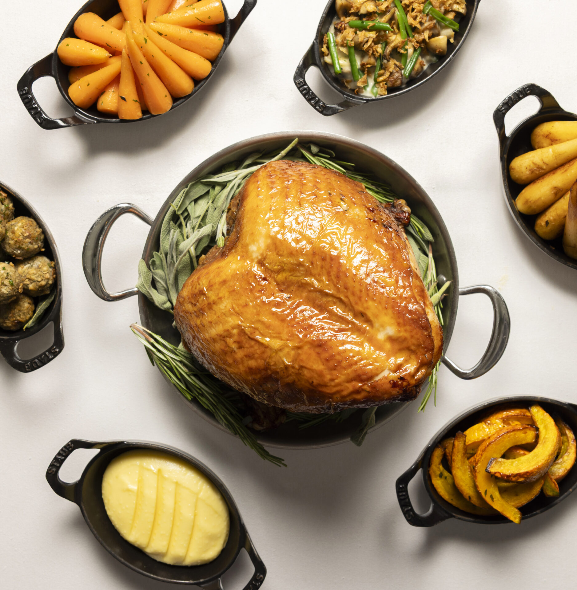 A roasted turkey sits on a bed of herbs in a round pan, surrounded by small dishes with carrots, green beans, potatoes, stuffing, mashed potatoes, and roasted squash on a white background.