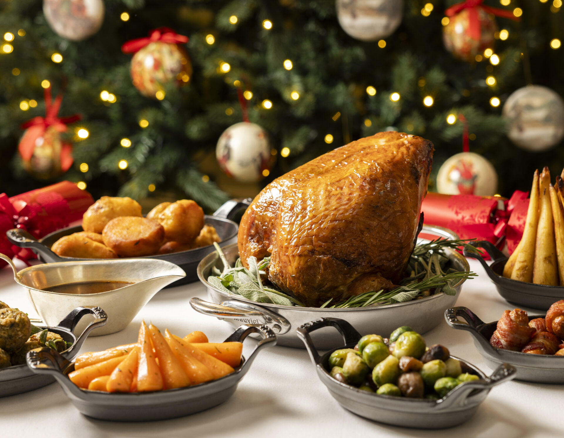 A festive table set with a roasted turkey, roast potatoes, carrots, Brussels sprouts, gravy, and parsnips, with a decorated Christmas tree and ornaments glowing in the background.