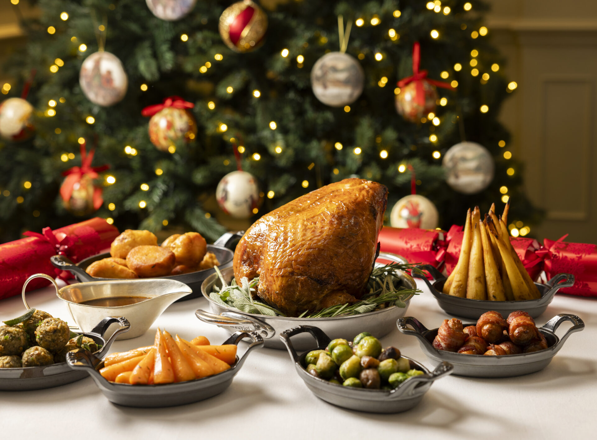 A festive table is set with a roasted turkey, vegetables, potatoes, gravy, and side dishes in front of a decorated Christmas tree with lights and ornaments.