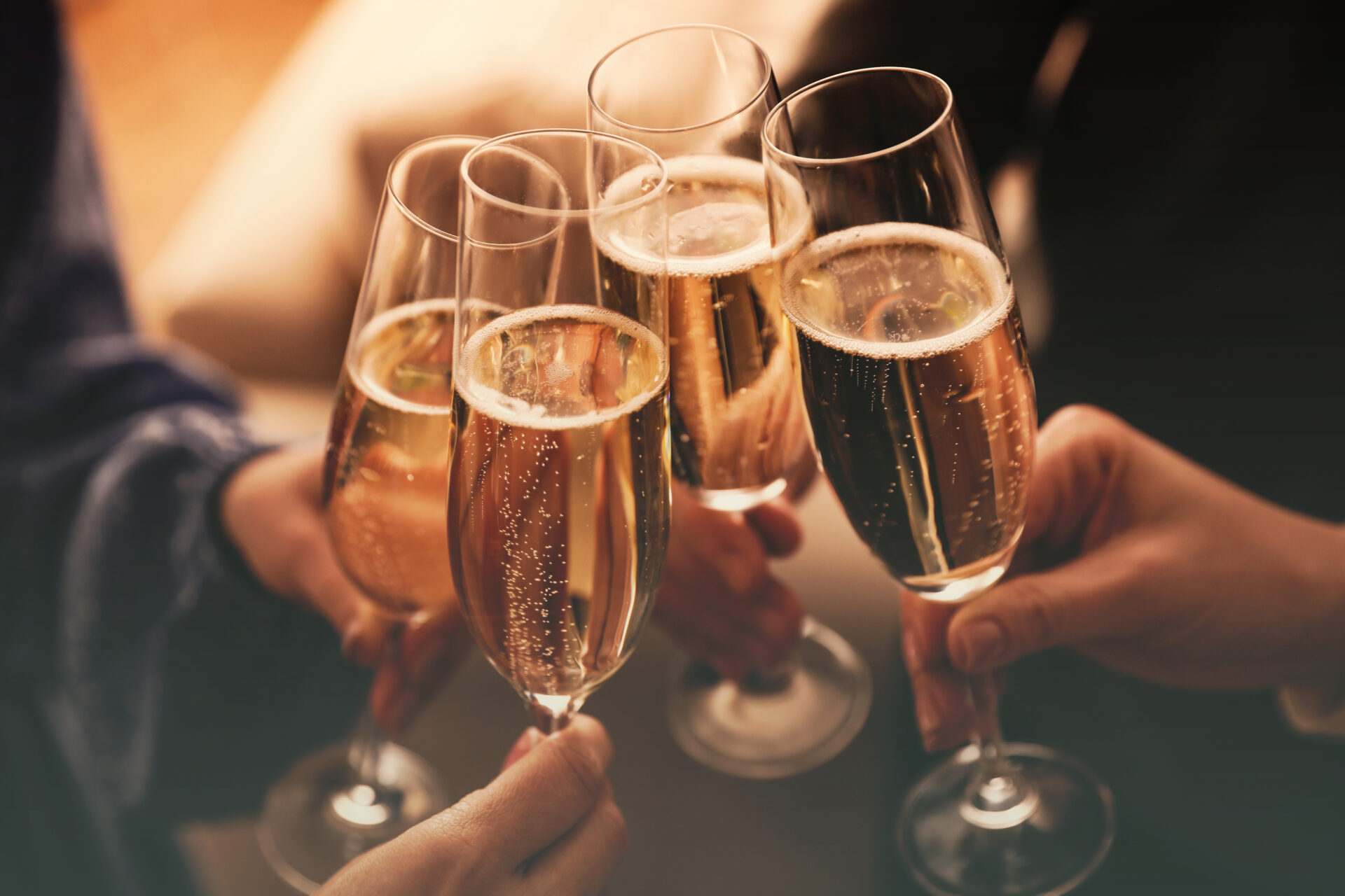 Four people clinking champagne glasses together in a toast, with sparkling wine and bubbles visible inside the glasses. The background is softly blurred.