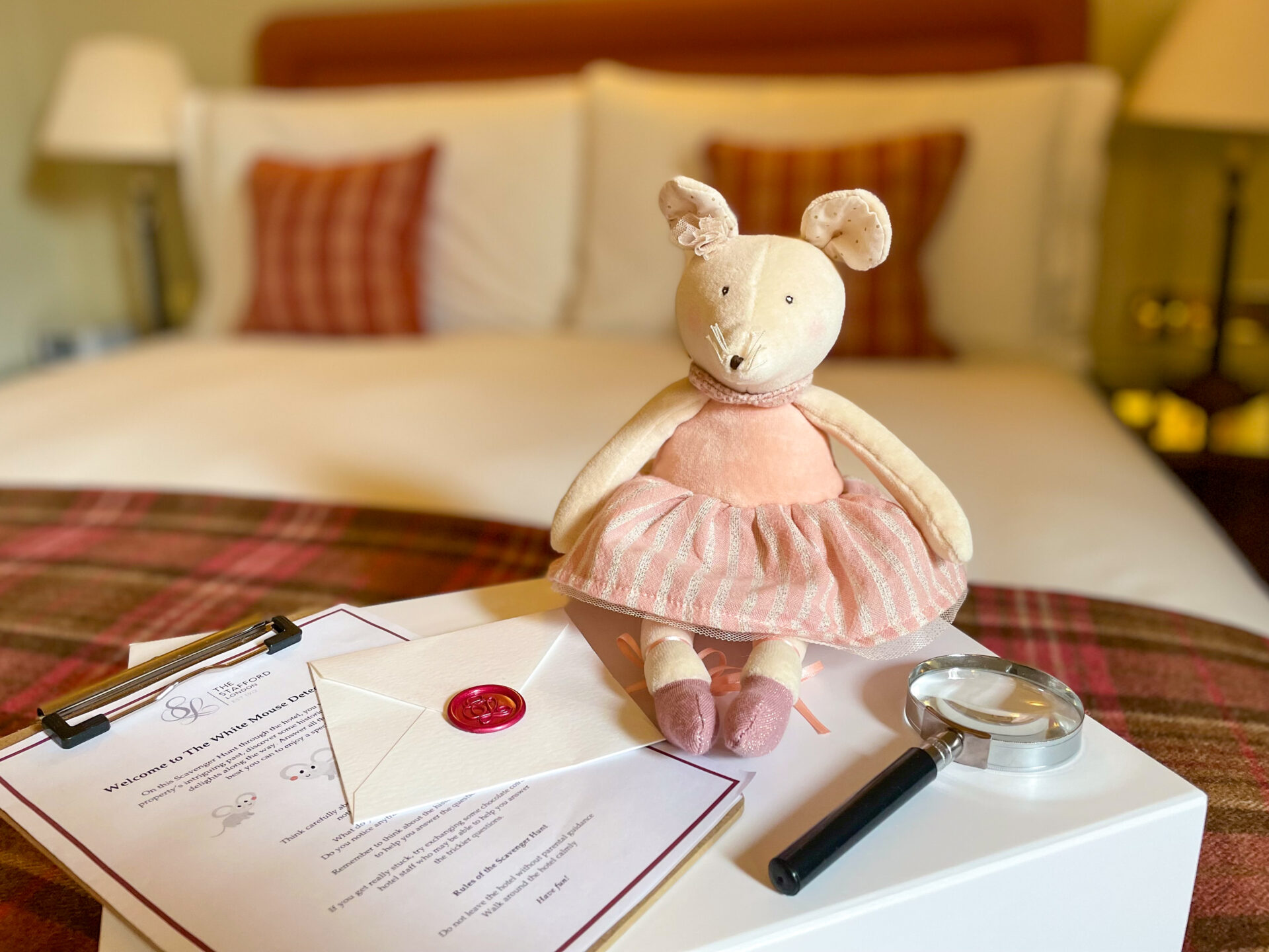 A plush mouse doll in a pink dress sits on a bed next to an envelope, a magnifying glass, papers, and a clipboard in a cozy, warmly-lit bedroom with plaid accents.