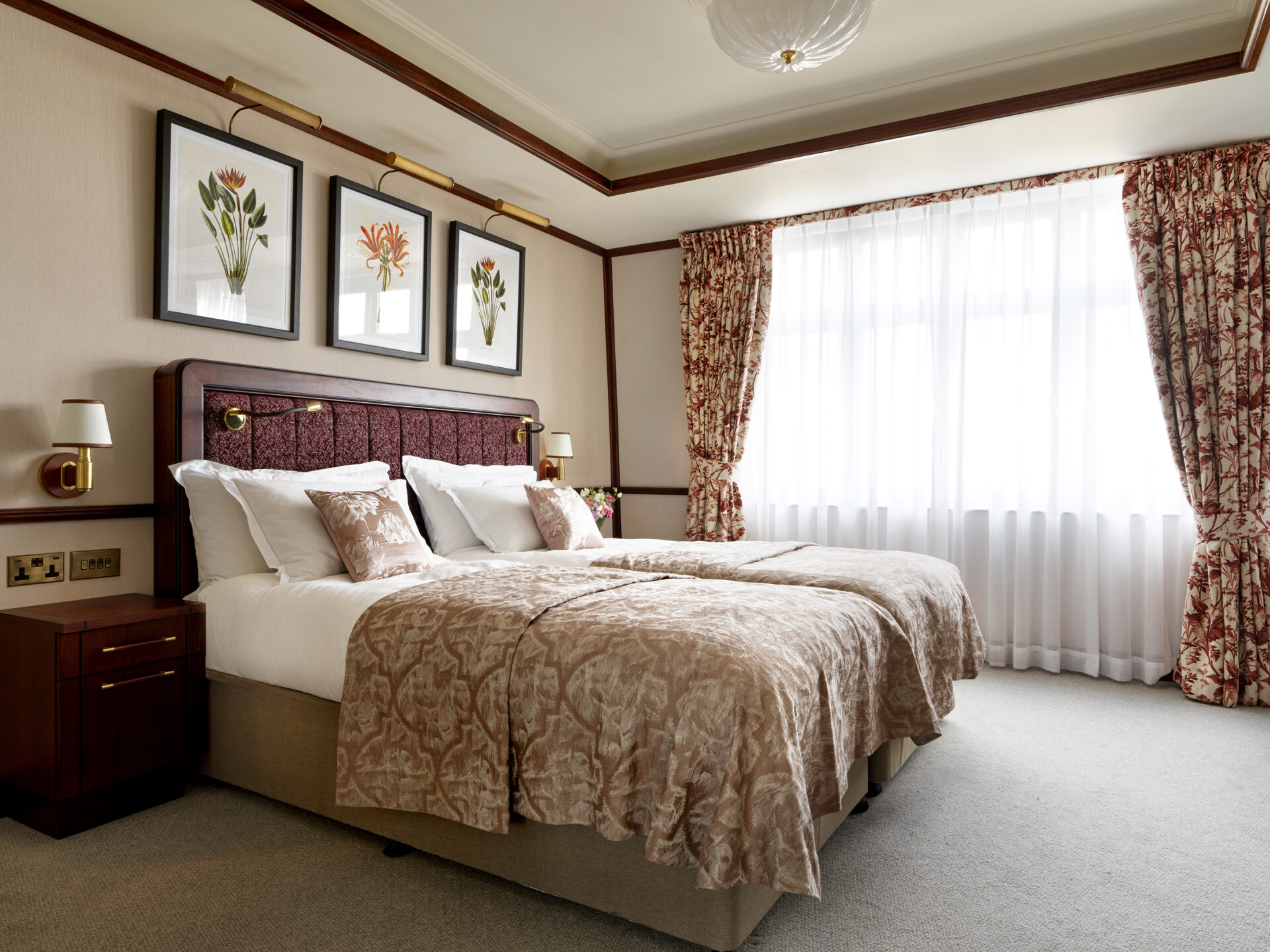 A well-lit hotel room with two twin beds pushed together, each with beige patterned bedding and white pillows. Decorative floral paintings hang above the headboard, and large windows with floral curtains brighten the space.